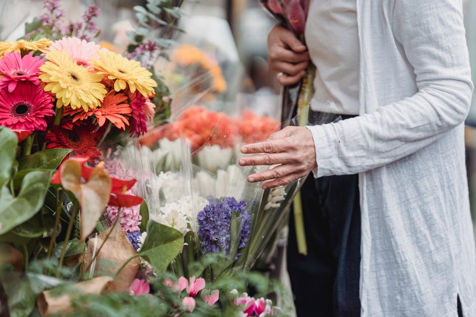 A person wearing a light grey coat with rolled-up sleeves is selecting a fresh flower bouquet at a florist stand. The bouquet includes bright gerbera daisies in shades of pink, yellow, and orange, arr