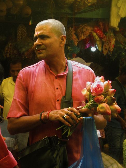 A man with a shaved head and medium skin tone, dressed in a salmon pink kurta, is holding a bouquet of pink and cream lotus flowers with green stems and unopened buds. He has a focused expression and 