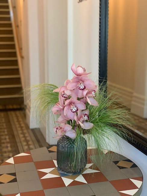 A tall floral arrangement featuring pink orchids with multiple blossoms on each stem, set in a dark, ribbed glass vase. The orchids have delicate, rounded petals with white centers and intricate purpl