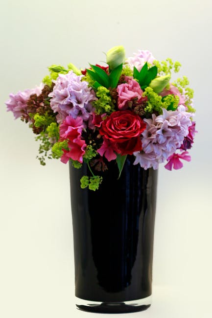 A vibrant floral bouquet featuring a mix of pink and purple hydrangeas, a single red rose, white tulips, and green foliage arranged in a tall, sleek black vase. The bouquet has a lush, full appearance