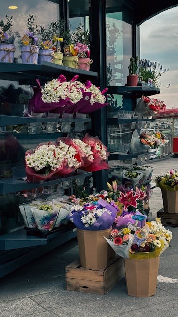 A display of various floral arrangements and bouquets inside a flower shop, with multiple shelves holding potted plants and wrapped bouquets. The flowers include white and pink chrysanthemums, purple