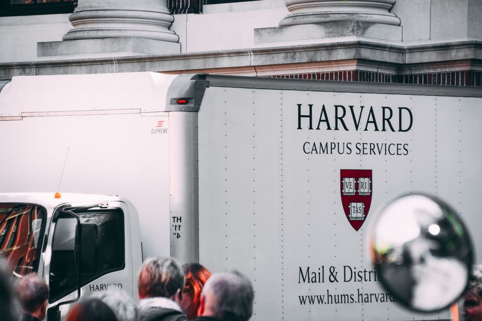 A close-up view of a large Harvard Campus Services delivery truck parked outside a classical building with stone columns, partially obscured by a crowd of people in the foreground. The truck is white