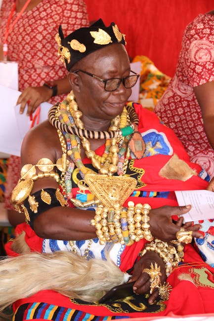 A woman dressed in traditional Maasai attire, adorned with multiple layers of large gold and beaded jewelry, including necklaces, bracelets, and earrings, wearing a black headpiece decorated with gold